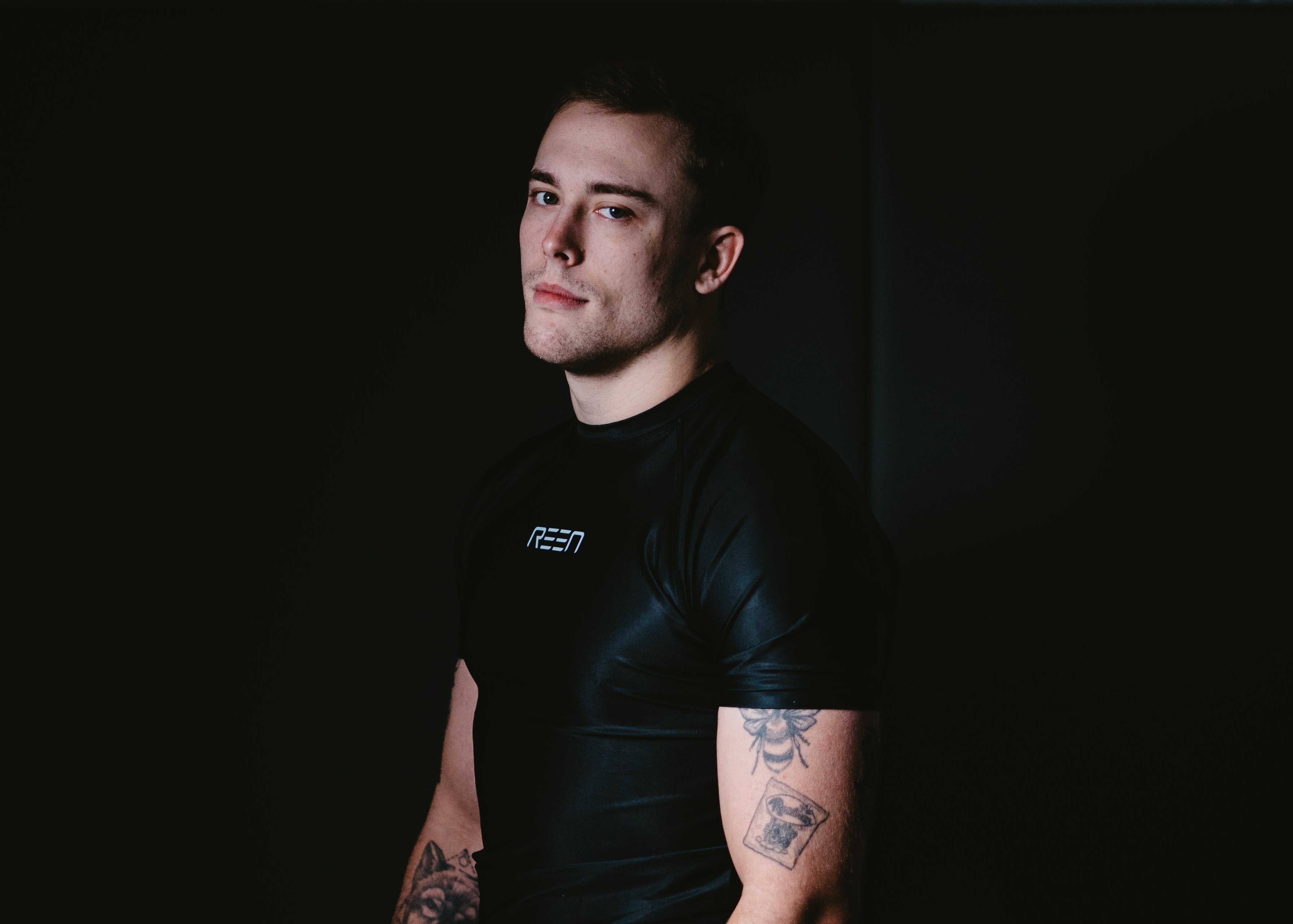 Man wearing a black t-shirt with visible tattoos sitting against a dark background